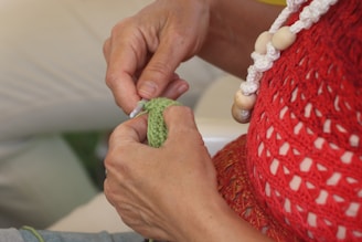 Hands holding a crochet hook working with green yarn, wearing a red crocheted top and a chunky necklace with white and wooden beads.