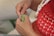 Hands holding a crochet hook working with green yarn, wearing a red crocheted top and a chunky necklace with white and wooden beads.
