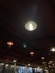 A series of hanging lights are suspended from the ceiling in a dimly lit room with a red roof structure. Some lights are illuminated, casting a warm glow, while others are off. The lighting creates a cozy and inviting atmosphere.