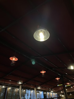 A series of hanging lights are suspended from the ceiling in a dimly lit room with a red roof structure. Some lights are illuminated, casting a warm glow, while others are off. The lighting creates a cozy and inviting atmosphere.