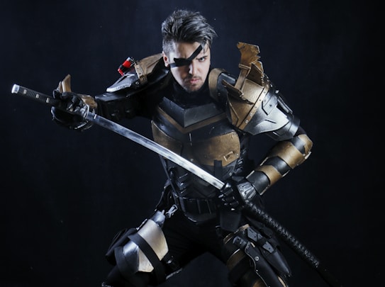 A person in a futuristic or fantasy-inspired armor is holding a long sword. The armor is predominantly metallic with gold and black accents, and features intricate detailing and sharp angles. The character has an intense expression and wears an eyepatch, adding to the warrior aesthetic.