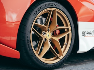 A close-up view of a car's rear wheel, featuring a prominent gold alloy rim with the brand logo at the center. The tire is a high-performance Michelin brand, complemented by a bright red brake caliper. The car's glossy red body contrasts with a white section that has a partially visible logo.