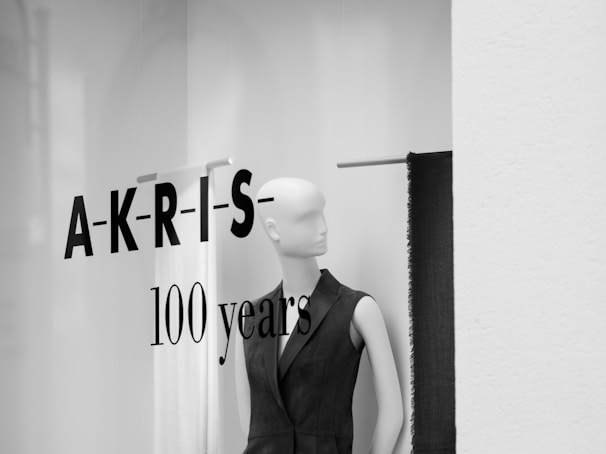 A mannequin dressed in a dark sleeveless outfit is displayed in a minimalist fashion store window. The words 'A-K-R-I-S- 100 years' are printed in bold letters on the glass, suggesting an anniversary celebration.
