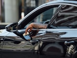 A tattooed arm rests on the window of a sleek black car, reflecting urban architecture in its glossy surface. The side mirror shows part of a person's face with sunglasses.