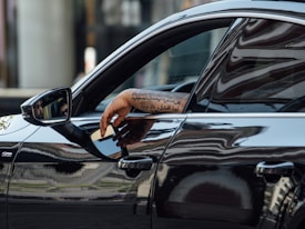 A tattooed arm rests on the window of a sleek black car, reflecting urban architecture in its glossy surface. The side mirror shows part of a person's face with sunglasses.