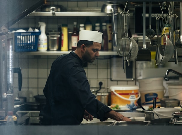 A chef wearing a white cap is working in a busy kitchen. There are various kitchen tools and utensils hanging above, and shelves are stocked with condiments and cleaning supplies. The atmosphere is bustling as the chef focuses on preparing food.