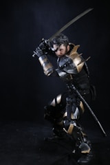 A person wearing elaborate medieval-style armor is posed in a crouching position while holding a sword. The armor is adorned with metallic elements and appears to be a combination of gold and silver colors. The dark background contrasts with the reflective surfaces of the armor, emphasizing the warrior-like stance.