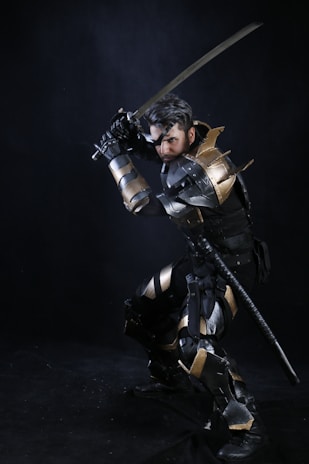 A person wearing elaborate medieval-style armor is posed in a crouching position while holding a sword. The armor is adorned with metallic elements and appears to be a combination of gold and silver colors. The dark background contrasts with the reflective surfaces of the armor, emphasizing the warrior-like stance.