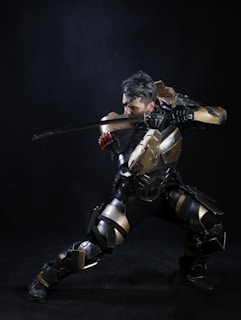 A person wearing a futuristic, armored costume poses with a sword raised in a dynamic, action-oriented stance. The armor is metallic, with gold and black tones, featuring intricate design elements. The background is dark, emphasizing the figure and the shiny reflections on the armor.