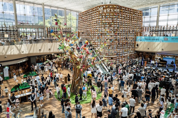 A large, bright indoor space filled with people. The area features a towering wall lined with numerous bookshelves, creating an extensive library atmosphere. In the center, there is an artistic installation resembling a tree adorned with colorful decorations and flowers. People are gathered around, engaging in various activities, some standing and others moving. The surroundings are well-lit with natural light coming through the large windows.