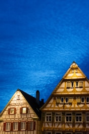 Two traditional timber-framed buildings are illuminated against a vibrant, deep blue evening sky. The structures feature intricate wooden detailing and numerous windows, some of which are lit from the inside.