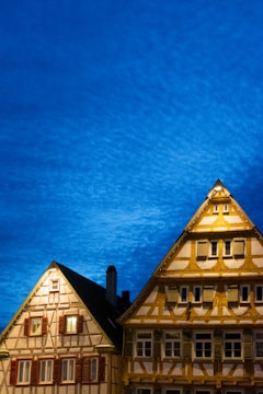 Two traditional timber-framed buildings are illuminated against a vibrant, deep blue evening sky. The structures feature intricate wooden detailing and numerous windows, some of which are lit from the inside.