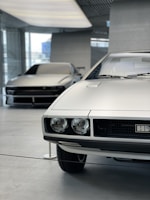 There are two cars in a modern, clean indoor setting. The car in the foreground has a boxy, classic design with round headlights and a silver exterior. The car in the background also has a sleek, silver exterior with a more modern, streamlined design. The floor is polished concrete, and there are large windows letting in natural light.