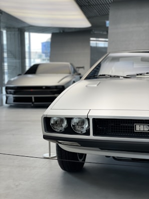 There are two cars in a modern, clean indoor setting. The car in the foreground has a boxy, classic design with round headlights and a silver exterior. The car in the background also has a sleek, silver exterior with a more modern, streamlined design. The floor is polished concrete, and there are large windows letting in natural light.