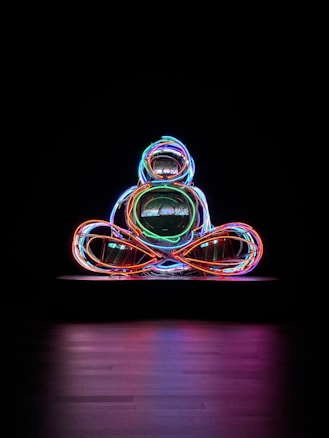 A vibrant and abstract sculpture made from intertwined neon lights in various colors is set against a dark background. The lights form a shape that suggests a seated figure or abstract design.