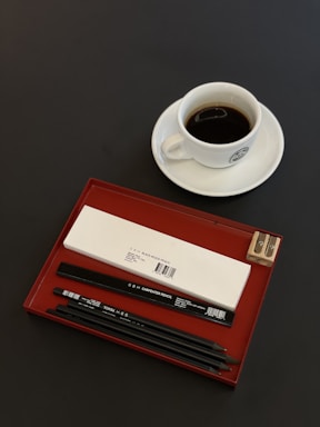 A sleek black pencil resting on a clean white desk beside a notebook and a cup of coffee.