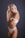A woman with long blonde hair is posing in white lingerie against a dark gray background. Her left hand is touching her hair while her right arm is crossed over her body. She has a solemn expression and there is a tattoo visible on her left arm near her shoulder.
