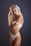 A woman with long blonde hair is posing in white lingerie against a dark gray background. Her left hand is touching her hair while her right arm is crossed over her body. She has a solemn expression and there is a tattoo visible on her left arm near her shoulder.