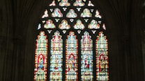 An elaborate stained glass window in a Gothic-style cathedral, featuring vibrant depictions of religious figures within tall, narrow panels. The intricate patterns and colorful artwork are framed by a dark stone architecture.
