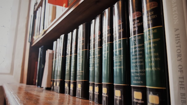 A row of large, dark green hardcover books is neatly arranged on a wooden shelf. The light from a nearby window illuminates the spines, which display gold lettering with book titles and volumes. Other books of varying sizes and colors fill the adjacent space on the shelves.