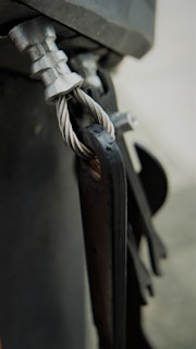 A close-up view of a metal cable secured with a clamp and attached to a metallic structure. The twisted cable is visible, showcasing its detailed texture and sheen. The components exhibit a rugged, industrial look.