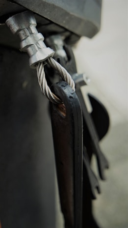 A close-up view of a metal cable secured with a clamp and attached to a metallic structure. The twisted cable is visible, showcasing its detailed texture and sheen. The components exhibit a rugged, industrial look.