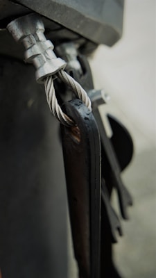 A close-up view of a metal cable secured with a clamp and attached to a metallic structure. The twisted cable is visible, showcasing its detailed texture and sheen. The components exhibit a rugged, industrial look.