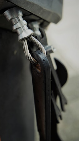 A close-up view of a metal cable secured with a clamp and attached to a metallic structure. The twisted cable is visible, showcasing its detailed texture and sheen. The components exhibit a rugged, industrial look.