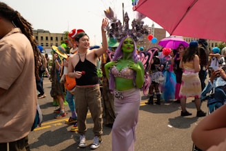 A lively outdoor event with people dressed in colorful and creative costumes, including a person with green body paint and a purple-themed outfit with butterfly wings, and another person wearing a red fish hat. There are vibrant accessories, including hats and umbrellas, while others in the background wear colorful wigs and elaborate outfits.