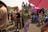 A lively outdoor event with people dressed in colorful and creative costumes, including a person with green body paint and a purple-themed outfit with butterfly wings, and another person wearing a red fish hat. There are vibrant accessories, including hats and umbrellas, while others in the background wear colorful wigs and elaborate outfits.