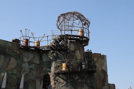 A structure made of corrugated metal panels and scaffolding, topped with a geodesic dome. Rusty barrels and arrow-like projections are visible, giving an industrial and post-apocalyptic look.