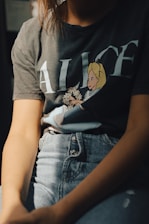 A person is wearing a t-shirt with an illustration of a cartoon character holding flowers. The t-shirt has the word 'Alice' printed on it. The person is also wearing high-waisted jeans with a button fly. The lighting casts shadows, creating an intimate, relaxed atmosphere.