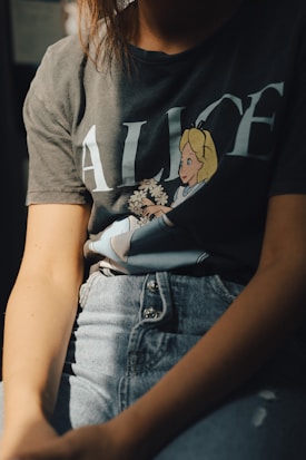 A person is wearing a t-shirt with an illustration of a cartoon character holding flowers. The t-shirt has the word 'Alice' printed on it. The person is also wearing high-waisted jeans with a button fly. The lighting casts shadows, creating an intimate, relaxed atmosphere.