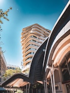 A modern urban setting featuring a tall, architecturally unique building with curved balconies and a smaller structure in the foreground with sweeping, wave-like architectural elements. The sky is clear with a deep blue tone, and there are some trees visible, adding a touch of greenery to the scene.