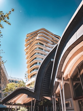 A modern urban setting featuring a tall, architecturally unique building with curved balconies and a smaller structure in the foreground with sweeping, wave-like architectural elements. The sky is clear with a deep blue tone, and there are some trees visible, adding a touch of greenery to the scene.