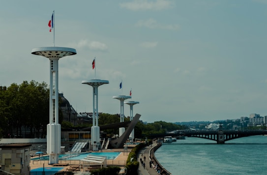 Large flagpoles with circular platforms and flags are positioned alongside a river. A pool with empty chairs and tables can be seen nearby. A bridge is visible in the background as well as a few people walking along the river pathway. Trees are on the opposite side of the flagpoles, and the sky is partly cloudy.