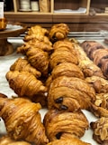 A display of freshly baked croissants arranged in rows on a marble counter. The pastries have a flaky, golden-brown crust, with some featuring chocolate fillings and others topped with almond slices. The background shows shelves with kitchen items.