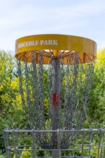 A disc golf basket is surrounded by chains and features a yellow band with 'Disc Golf Park' written on it. The structure is set against a backdrop of vibrant green foliage and yellow flowers, with a clear blue sky visible above.