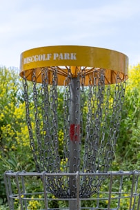 A disc golf basket is surrounded by chains and features a yellow band with 'Disc Golf Park' written on it. The structure is set against a backdrop of vibrant green foliage and yellow flowers, with a clear blue sky visible above.