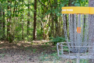 A disc golf basket stands in a wooded area with sunlight filtering through the trees. The basket, labeled with 'GOLF PARK,' is made of metal chains and a cage to catch discs. The surrounding area is filled with lush greenery and dense foliage, creating a peaceful, natural setting.