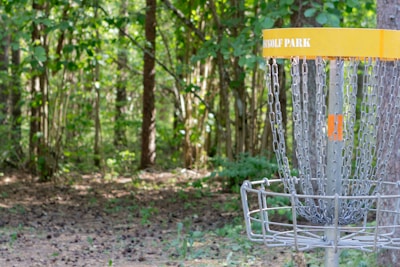 A disc golf basket stands in a wooded area with sunlight filtering through the trees. The basket, labeled with 'GOLF PARK,' is made of metal chains and a cage to catch discs. The surrounding area is filled with lush greenery and dense foliage, creating a peaceful, natural setting.