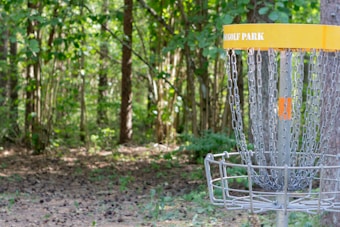 A disc golf basket stands in a wooded area with sunlight filtering through the trees. The basket, labeled with 'GOLF PARK,' is made of metal chains and a cage to catch discs. The surrounding area is filled with lush greenery and dense foliage, creating a peaceful, natural setting.