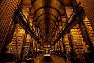 A grand, historic library hall with towering shelves filled with numerous books. The architecture features high, arching ceilings and dark wood paneling. Busts of historical figures line the central aisle, lending an air of scholarly gravitas. Warm, ambient lighting enhances the richness of the wood and creates a cozy yet awe-inspiring atmosphere.
