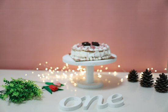 A cake with whipped cream and chocolate cookies sits on a white stand surrounded by soft, glowing fairy lights. Greenery and a tulip are laid on a white surface next to wooden pine cones. The word 'one' is prominently displayed in white.