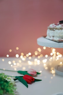 A pastel-colored backdrop features a cake adorned with decorative toppings on a white stand. Nearby, artificial tulips rest on the surface, with blurred fairy lights creating a warm ambiance.