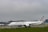 An airplane with Air France livery is on the ground at an airport. The aircraft is situated on the taxiway with its engines visible. In the background, an airport terminal with multiple buildings, including an air traffic control tower, and other airplanes can be seen. The sky appears overcast, and there is green grass in the foreground.