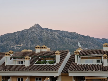 A range of rugged, rocky mountains is visible in the background, set against a soft, pale pink sky at dusk or dawn. In the foreground, there are several traditional Spanish-style houses with terracotta-tiled roofs, white walls, and wooden pergolas. A satellite dish is mounted on one of the roof corners.