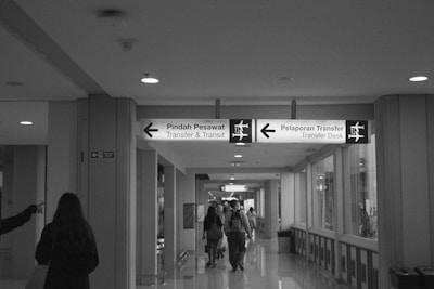 A corridor in an airport with signage showing directions for transfer and transit. There are people walking along the corridor, and the signage is in both Indonesian and English, directing passengers to 'Transfer & Transit' and 'Transfer Desk'. The corridor is well-lit with overhead lights, and there are windows on one side along with wall panels on the other.