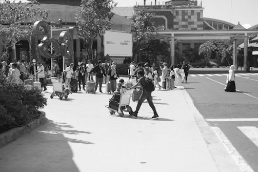A busy outdoor scene at an airport or transit hub with a large group of people, many with luggage and carts, moving across a paved area. Buildings and landscaping are visible in the background, with people appearing to be in transit or awaiting transport.