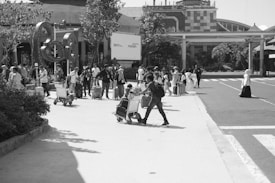 A busy outdoor scene at an airport or transit hub with a large group of people, many with luggage and carts, moving across a paved area. Buildings and landscaping are visible in the background, with people appearing to be in transit or awaiting transport.